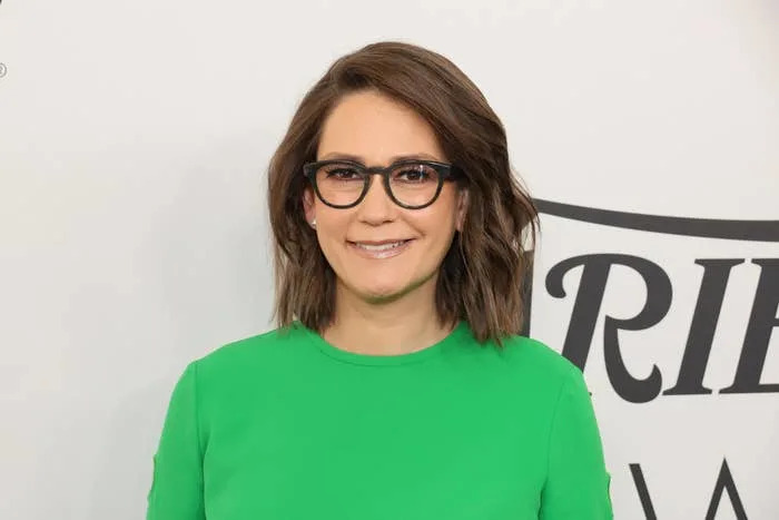 Person wearing glasses and a green dress at a public event. Neutral background