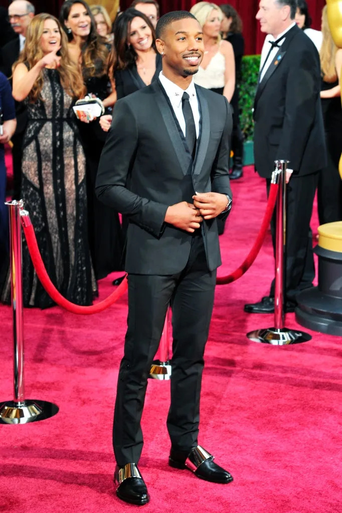 Person in a sleek suit smiles on the red carpet, surrounded by photographers and onlookers, showcasing an elegant and polished look