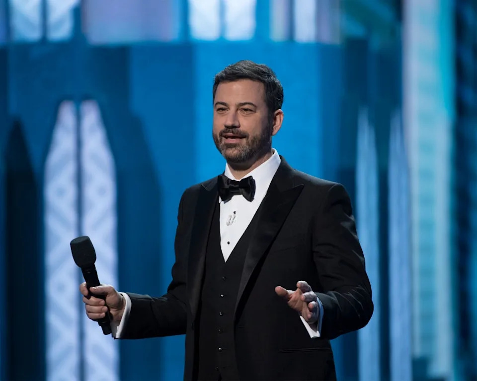 A person in a tuxedo stands on stage holding a microphone, appearing to speak during a formal event