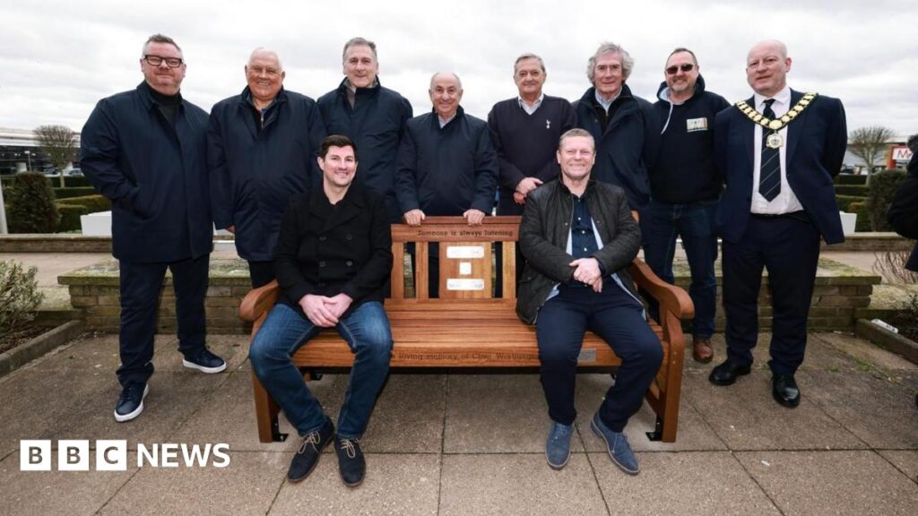 Several people gather around a brown wooden bench. Eight people are stood behind it, with two people sat on the bench. On the bench there are several plaques, and above them is some black writing which says "someone is always listening".