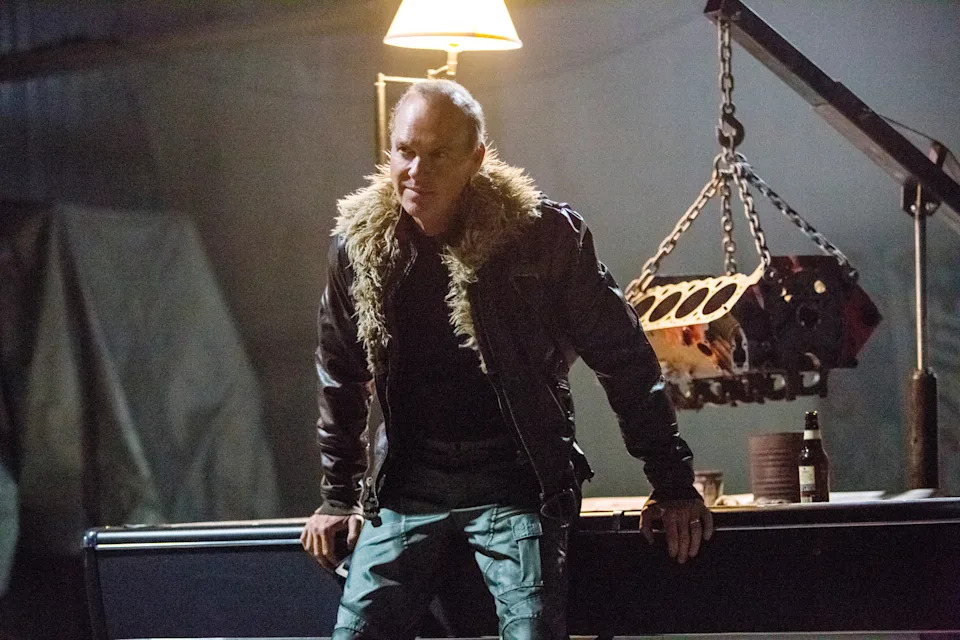 Man in a leather jacket with a fur collar in a dimly lit industrial setting, standing by a table with a lamp and engine parts