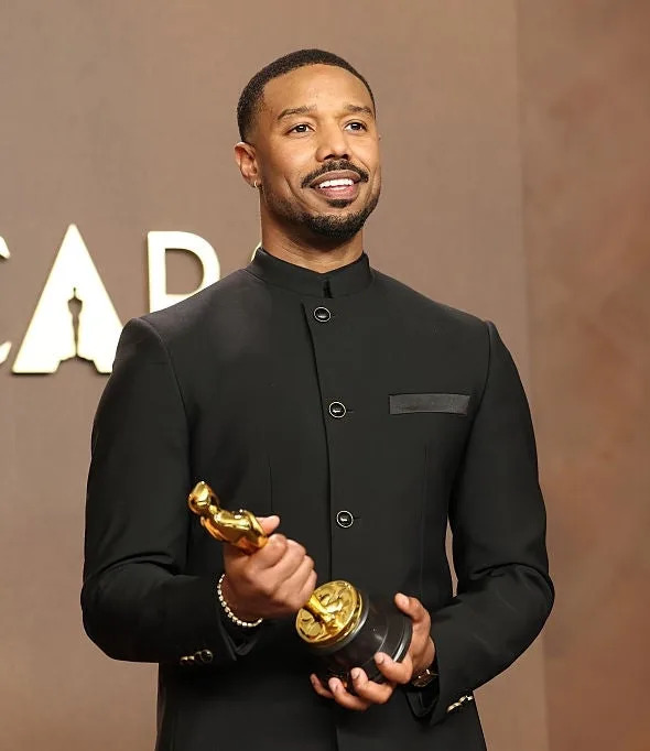 A person stands on the red carpet holding an Oscar, wearing a sleek, buttoned-up black suit