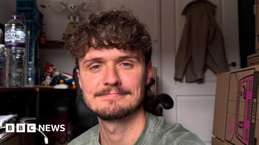 A selfie photograph of Ben Cole Edwards, who is 30 years old, has brown curly hair, a brown beard, and blue eyes. Behind him, a coat can be seen on the hook on his back door, and a plastic water bottle can be seen on a desk behind him.