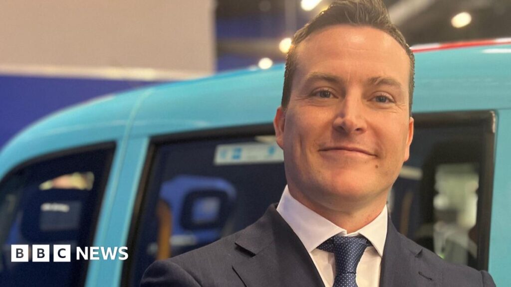 James McMurdock is in a navy suit and blue patterned tie, with what looks like a Hackney cab behind him, painted the sky blue colour of Reform UK.