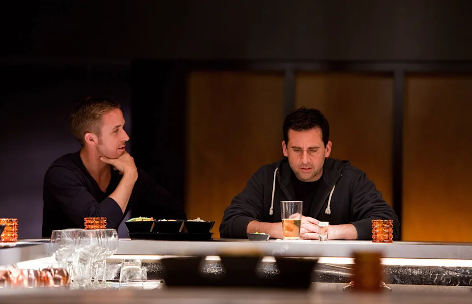 Two men sit at a bar engaged in conversation, one in casual attire with a drink. The setting suggests a reflective or intimate moment