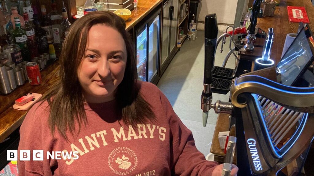 Megan stands behind the bar, smiling up at the camera. Behind her are bottles of alcohol and she stands next to the handles to pull pints. She has shoulder length brown hair and wears a salmon coloured hoodie with white writing on.