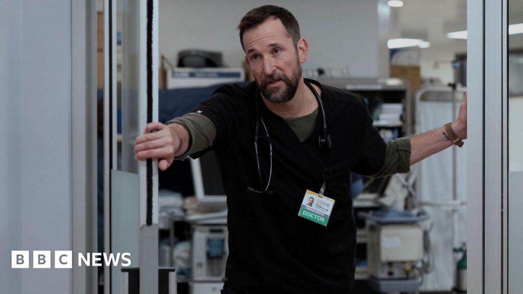 Dr. Michael “Robby” Robinavitch (Noah Wyle) looking exhausted, wearing black scrubs and holding a door open