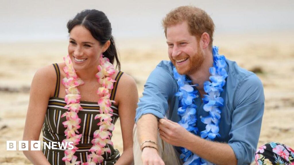Meghan Markle, wearing a striped black, white and brown dress, sitting next to Prince Harry, wearing a blue collared shirt with sleeves rolled up. Both are smiling and wearing garlands of flowers around their necks.