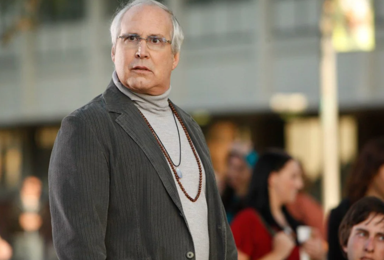 An older man with gray hair and glasses, wearing a gray turtleneck, striped blazer, and a beaded necklace, stands outdoors looking surprised. Blurry people and a building are visible in the background.