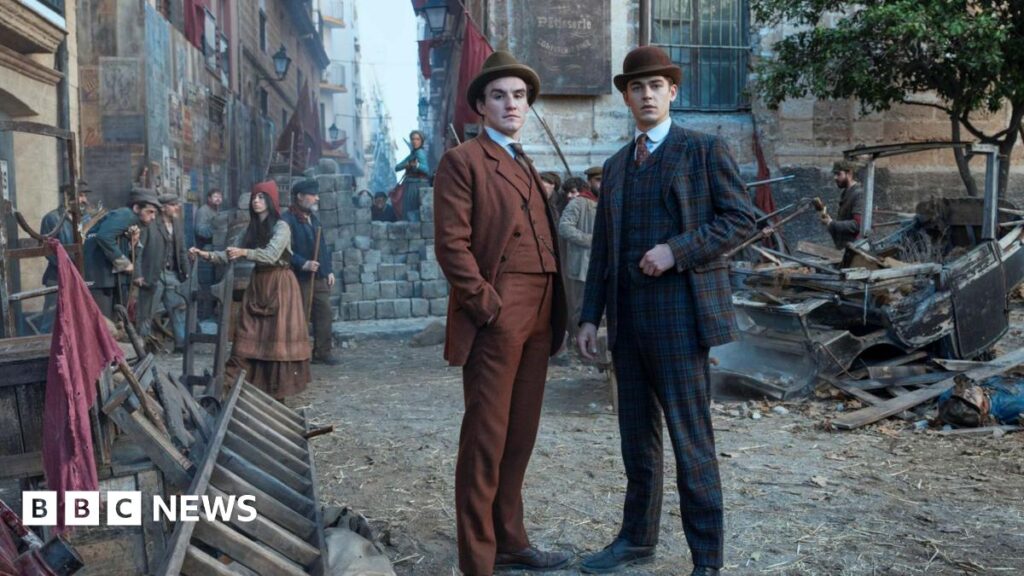 Young Sherlock stands with another man in an orange suit in a street made to look like a Victorian era London street. He wears a checked suit and a bowler hat