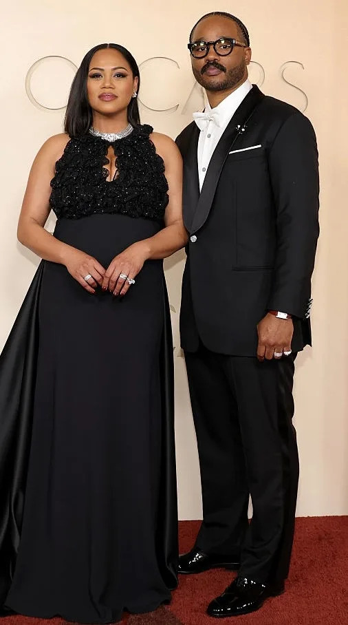 Two individuals stand on a red carpet; one wears an elegant black gown, the other a classic tuxedo