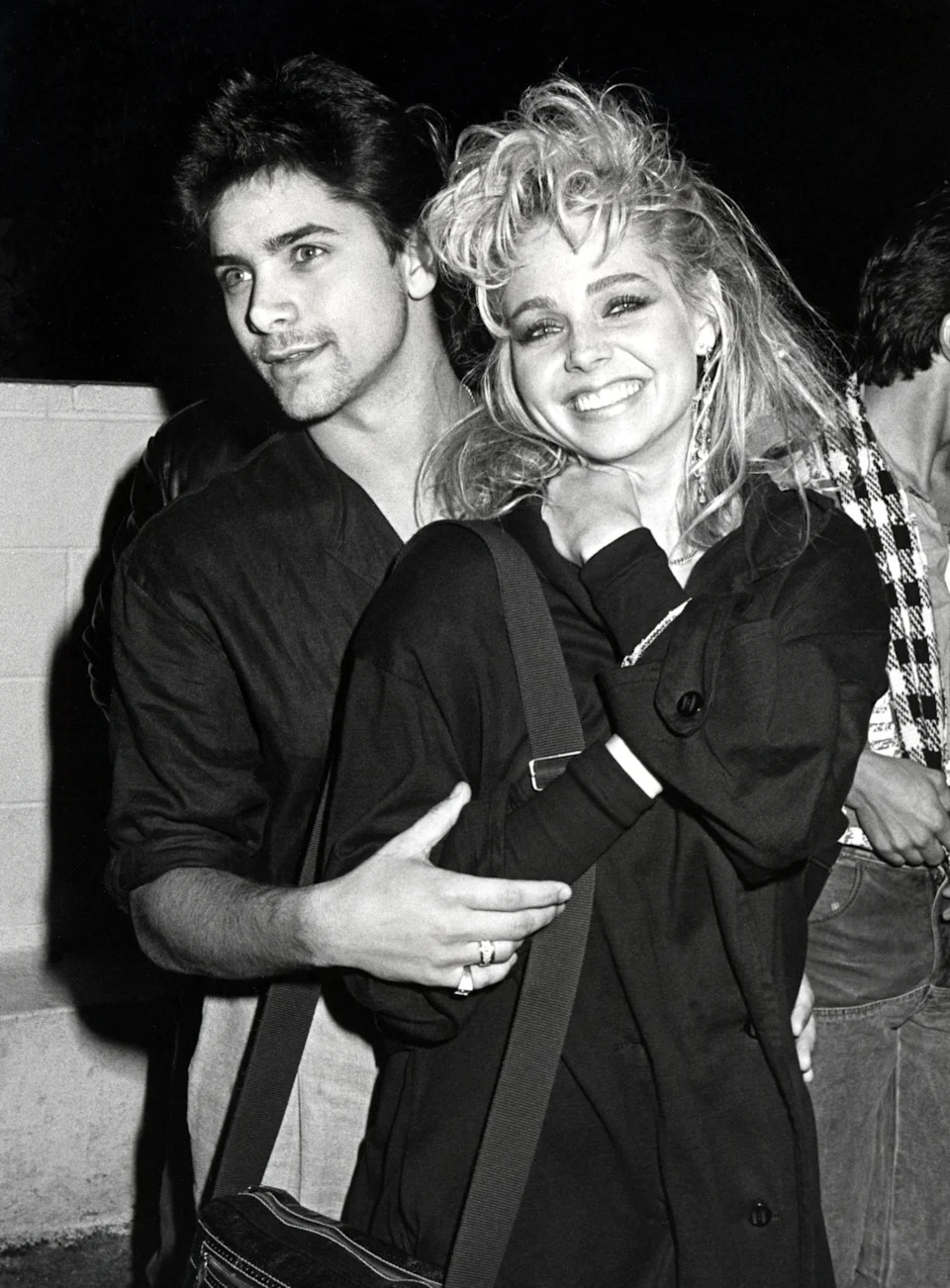 A smiling couple in 1980s fashion with big hair. The man wears a casual jacket, and the woman has a draped coat over her shoulders