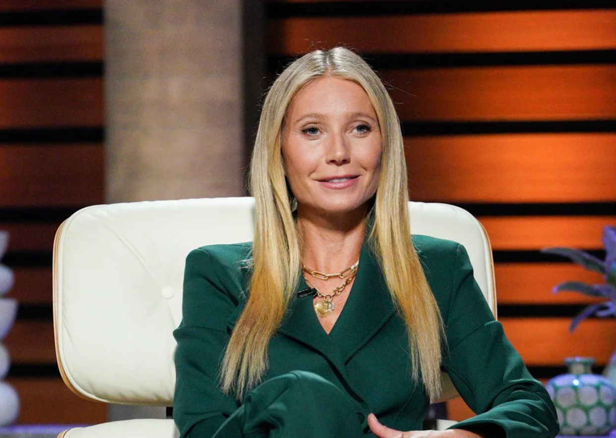 A woman with long blonde hair sits on a white chair, wearing a dark green suit and gold necklaces, against a background with warm-colored horizontal panels.