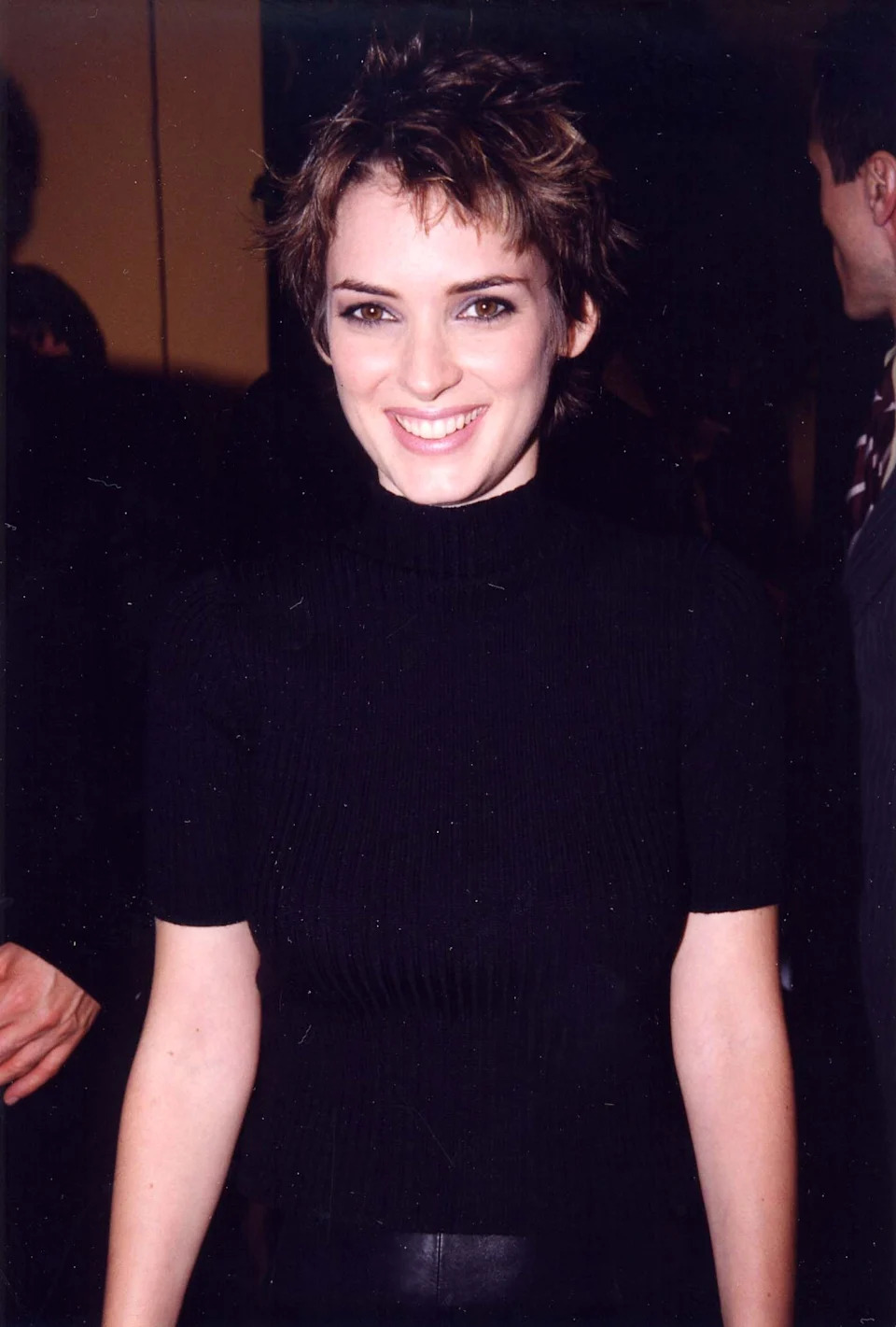 Winona Ryder smiling, wearing a fitted, ribbed black turtleneck. Short, tousled hair style. Indoor setting