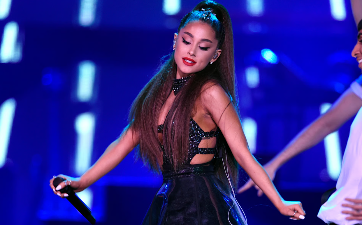 A woman with long brown hair in a high ponytail performs on stage wearing a sparkly black outfit. She holds a microphone and is smiling with her eyes closed, colorful stage lights glowing behind her.