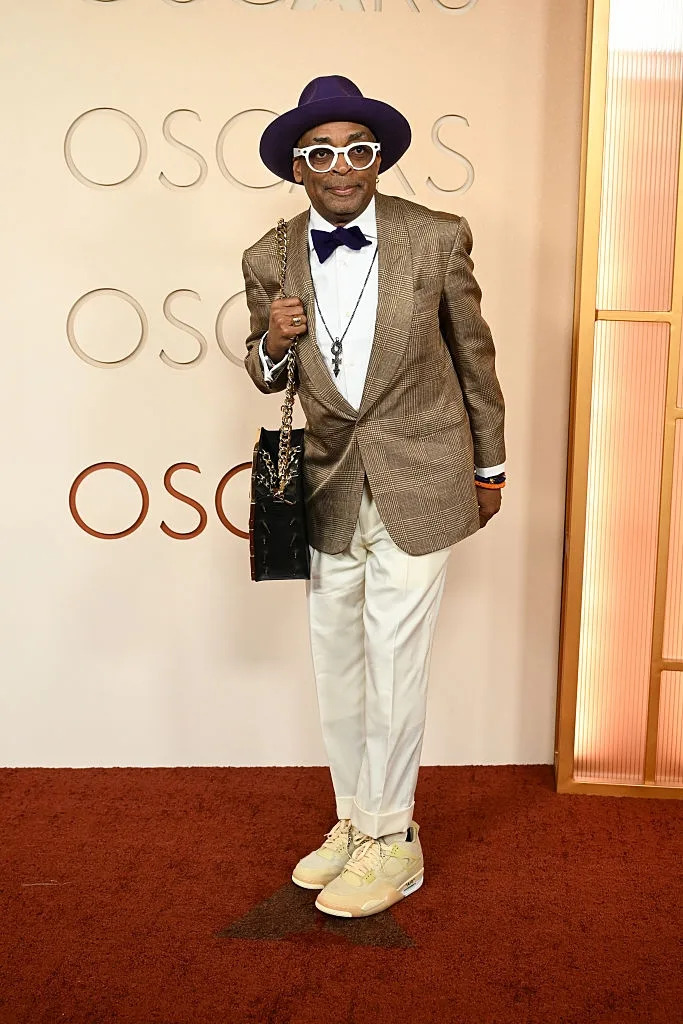Person in a brown plaid suit, purple hat, and bow tie on a red carpet, holding a bag. They're wearing white shoes and glasses