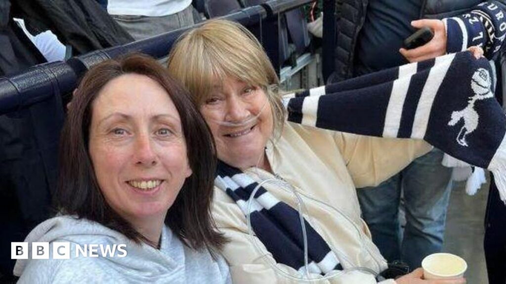 Two women sat in a football stadium wearing football scarves.