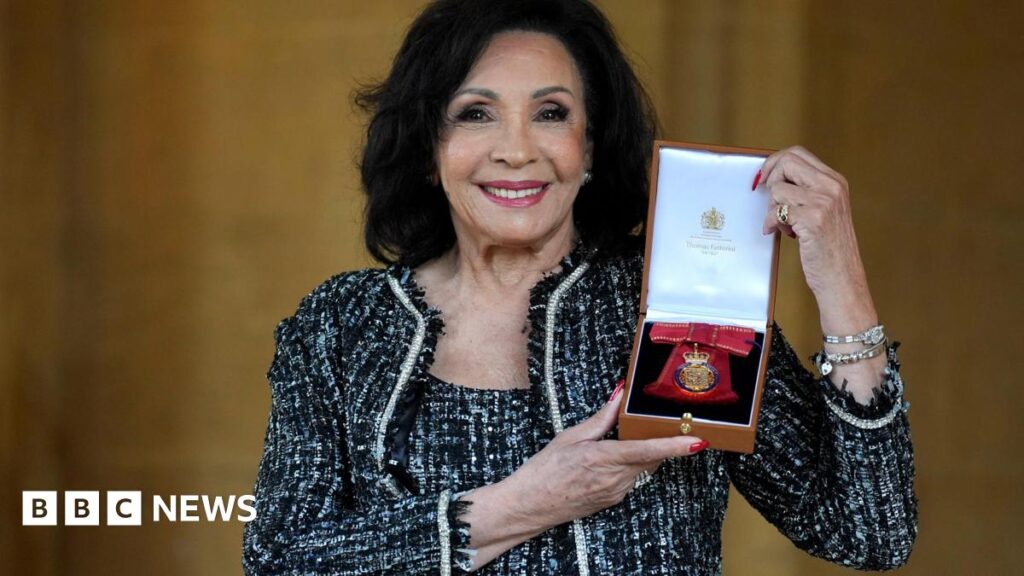 The image shows Dame Shirley Bassey holding her Companion of Honour award. She is wearing a black and white dress and matching jacket.