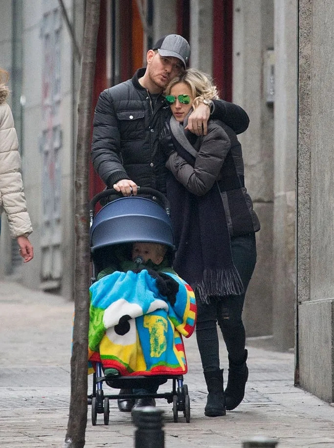 A couple walks on a city street embracing, pushing a stroller with a child under a colorful blanket