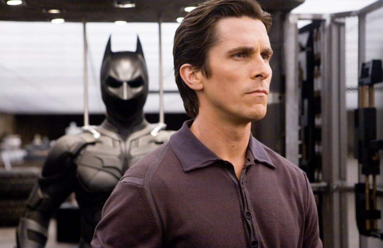 A man with short brown hair wearing a dark collared shirt stands in front of a Batman suit displayed on a mannequin in a modern, industrial setting.