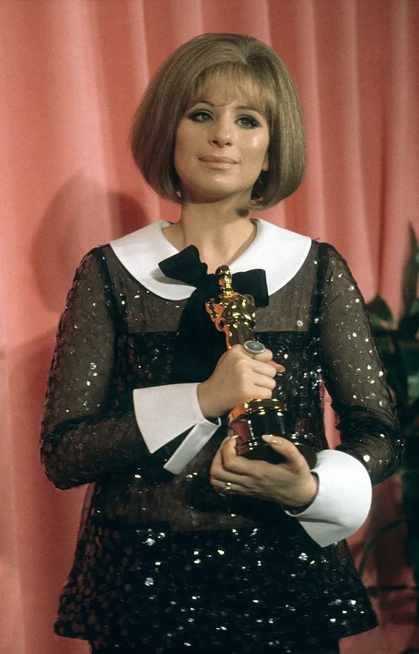 A person in a sheer, sequined dress with white collar and cuffs holds an award statue on a stage