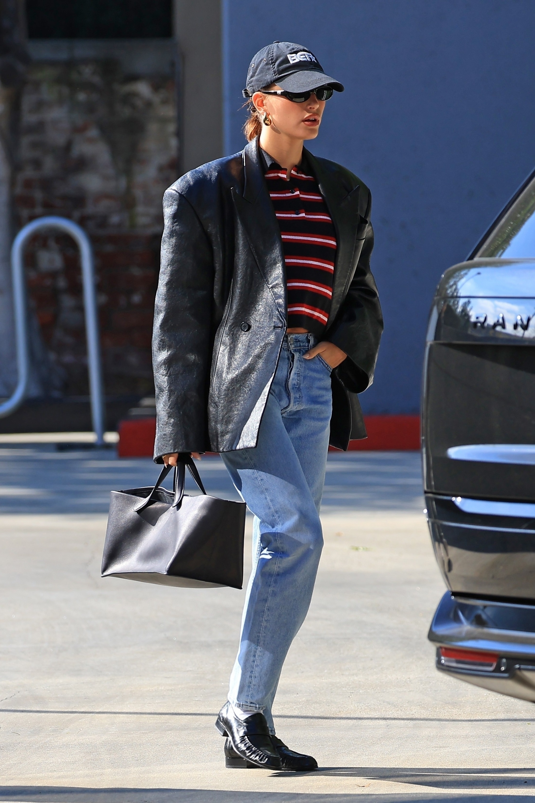 Hailey Bieber wears a leather jacket, striped top, blue jeans, black loafers and carries The Row Idaho bag