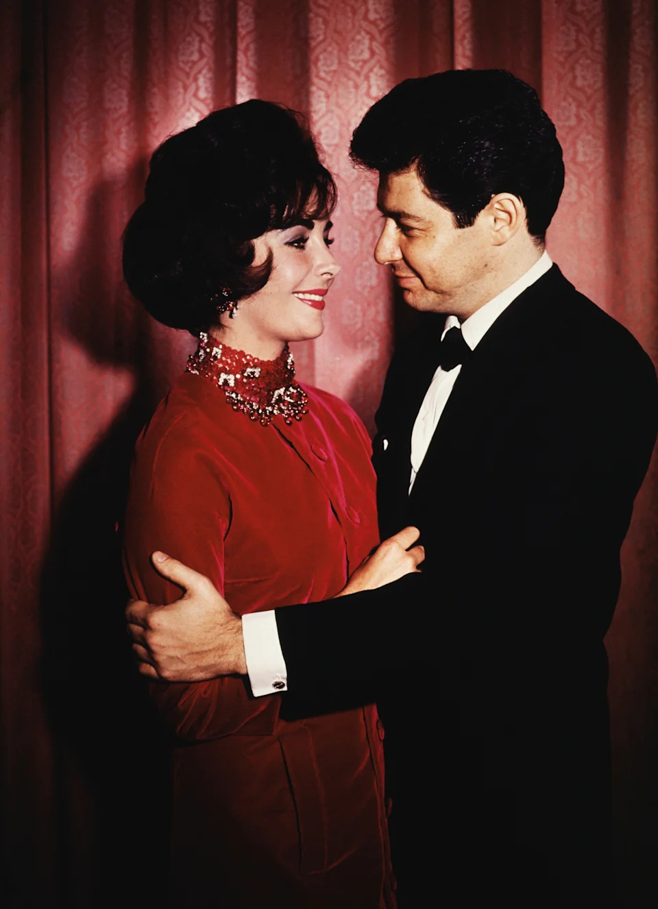 A couple dressed in elegant evening attire share an intimate moment, gazing into each other's eyes in front of a curtain backdrop