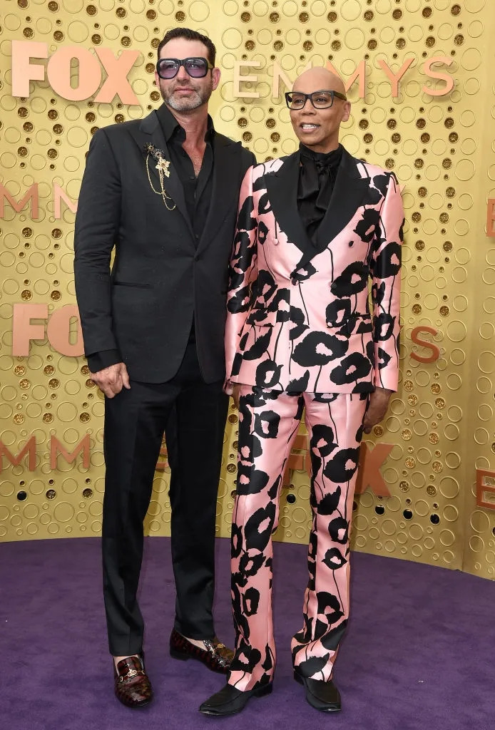 Two people on a red carpet. One in a black suit with a unique brooch, the other in a floral-patterned suit. Event backdrop indicates an awards show