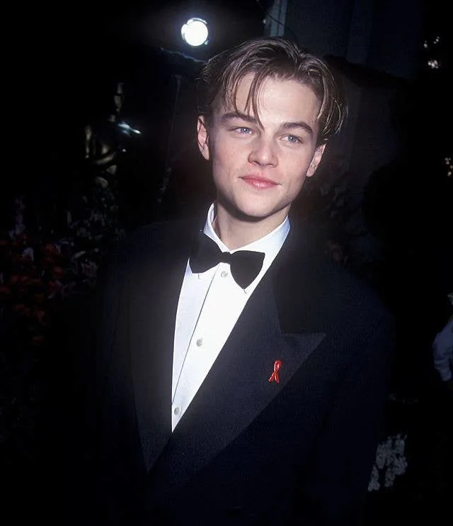A person in a tuxedo with a bow tie and a red ribbon, standing on a dimly lit red carpet