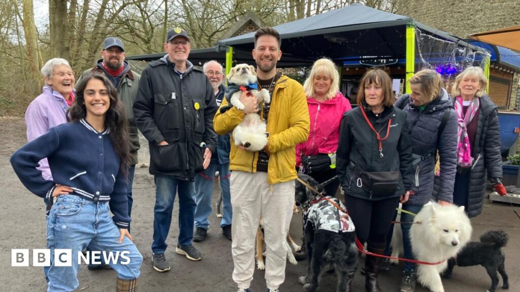A group of people stand outside, in a park with their dogs. The coffee stand is in the background, with an awning for people to sit underneath.
