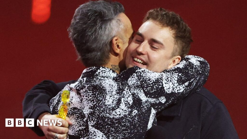 Robbie Williams has short grey hair and a black and white patterned formal jacket. He is embracing Sam Fender who has short brown hair and is holding a Brit Award. He is wearing a dark jacket. Olivia Dean is standing next to them with long dark hair and wearing a patterned black dress.