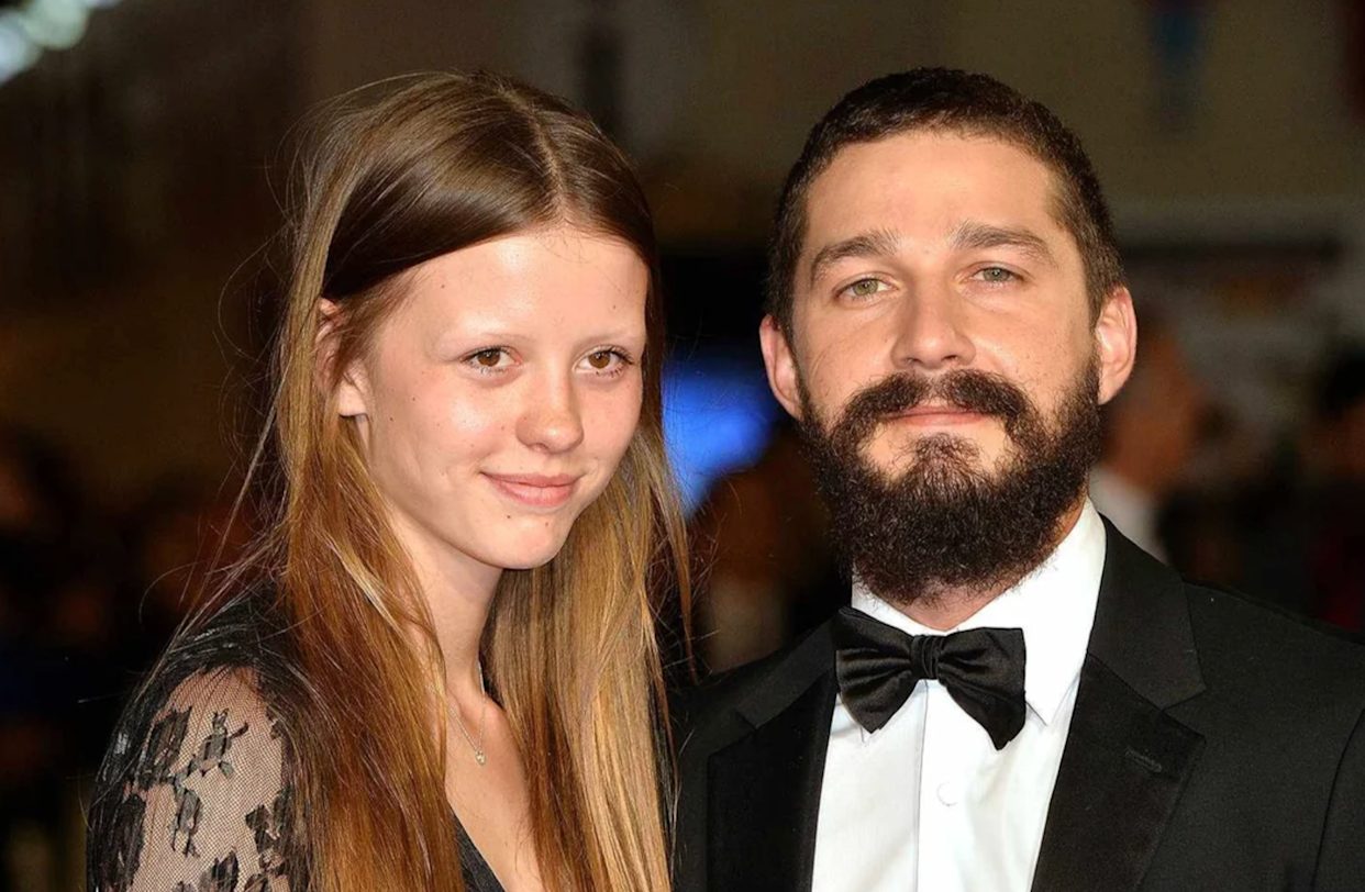 A young woman with long brown hair wearing a black lace dress stands next to a man with a full beard and mustache, dressed in a black tuxedo and bow tie, both smiling at an event with a blurred crowd in the background.