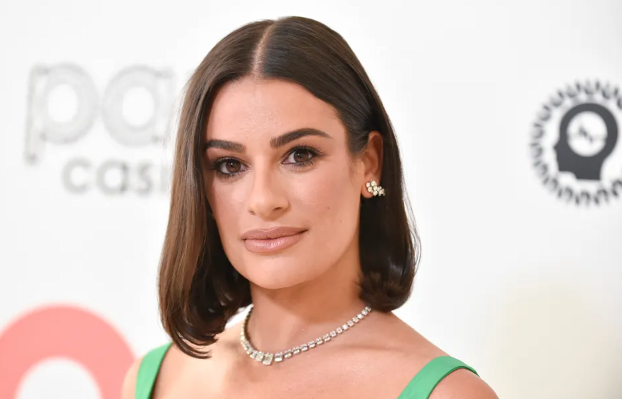 A woman with shoulder-length dark brown hair, wearing a green dress, diamond necklace, and pearl earrings, poses in front of a white background with logos.
