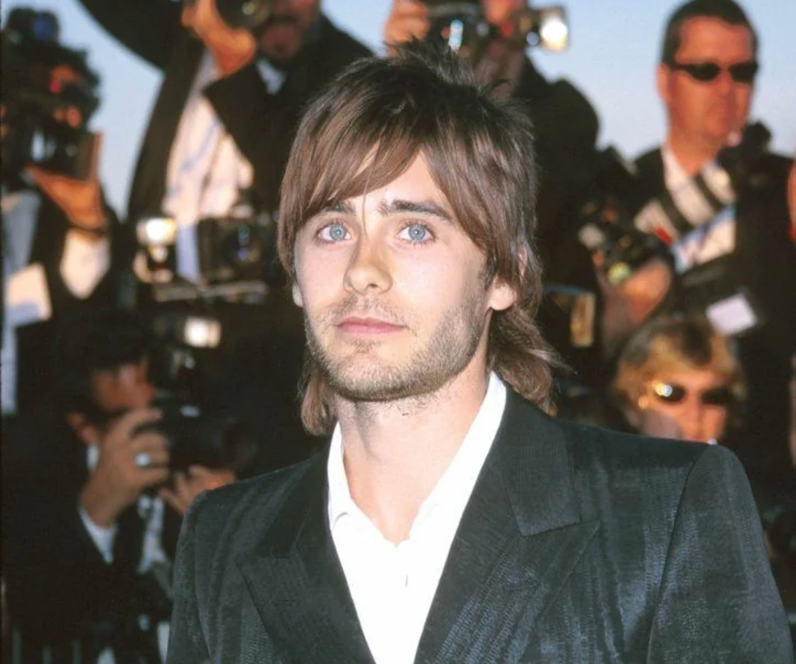 A man with medium-length brown hair and a trimmed beard wears a dark suit and stands in front of photographers at an event, with bright camera flashes and blurred people in the background.