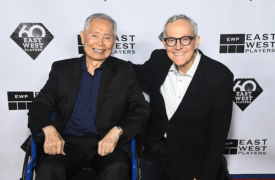 Two men smiling at an event; one seated in a wheelchair, both in suits, in front of an East West Players backdrop