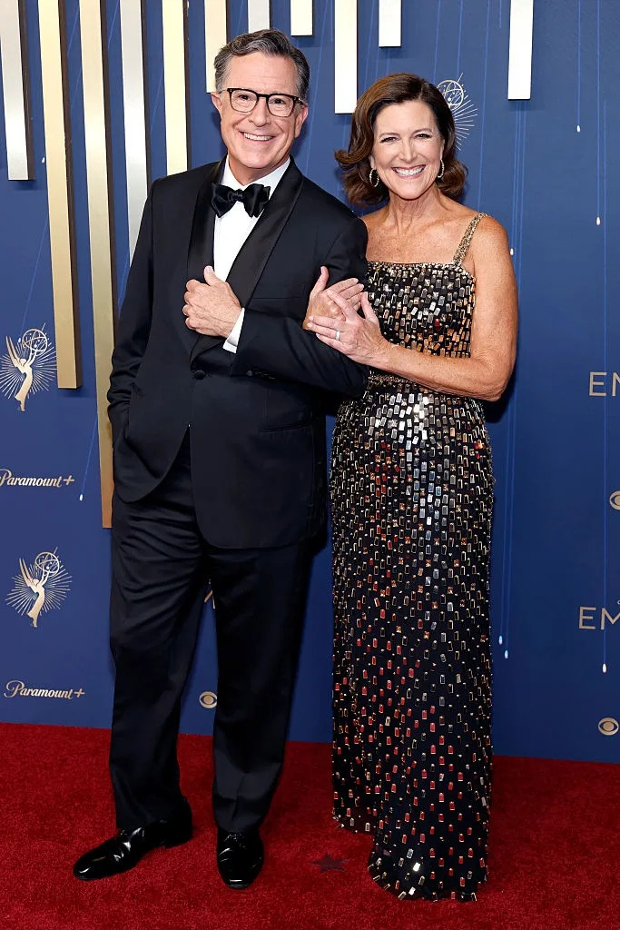 A couple poses on the red carpet; man in a classic tuxedo, woman in a sequined evening gown, both smiling and linking arms