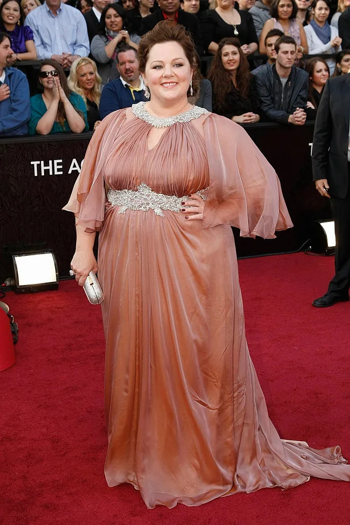 A person in an elegant, flowing gown poses confidently on the red carpet at a celebrity event, holding a small clutch bag
