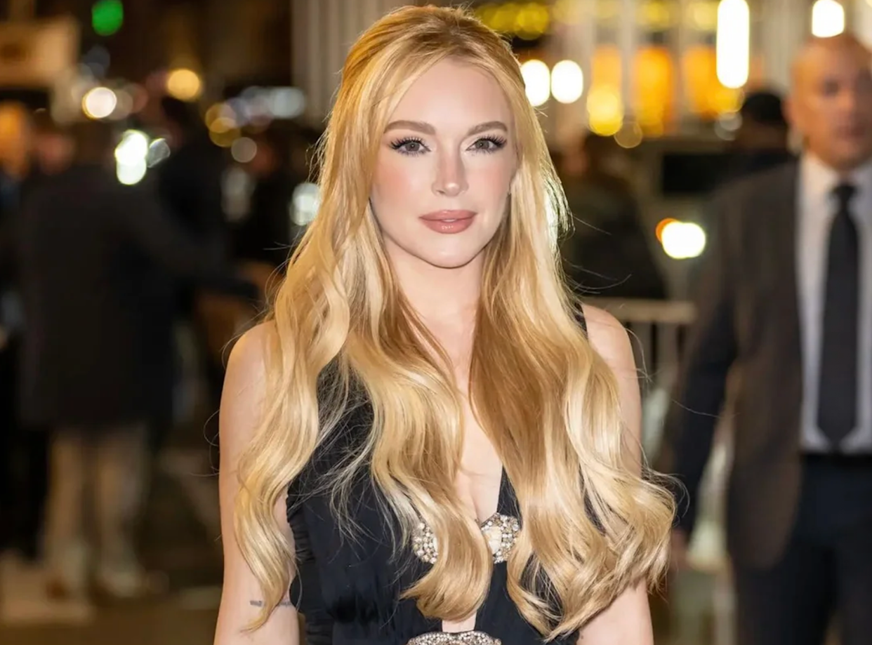 A woman with long, wavy blonde hair wearing a sleeveless black dress stands outdoors at night, with blurred people and city lights in the background.