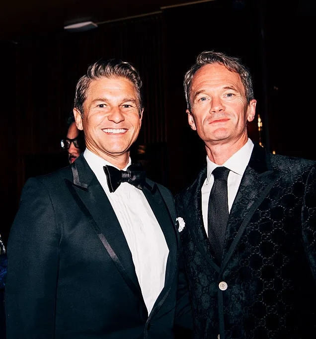 Two men in tuxedos at a formal event, smiling at the camera, with others in elegant attire in the background