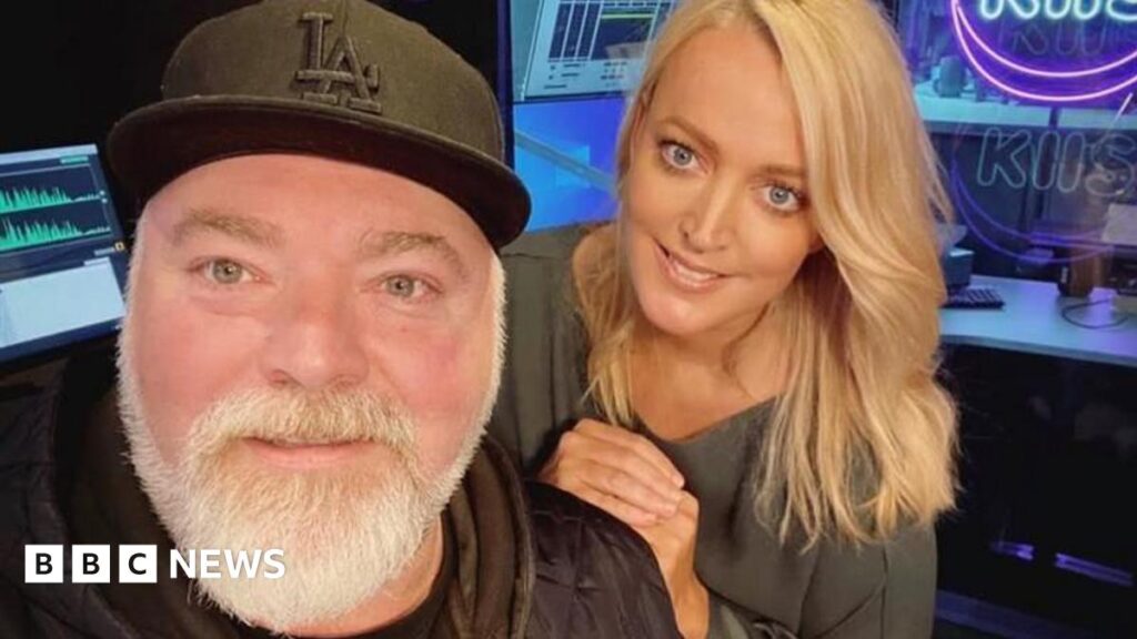 A middle aged man with a black baseball cap and grey beard and a blonde-haired younger woman smile at the camera