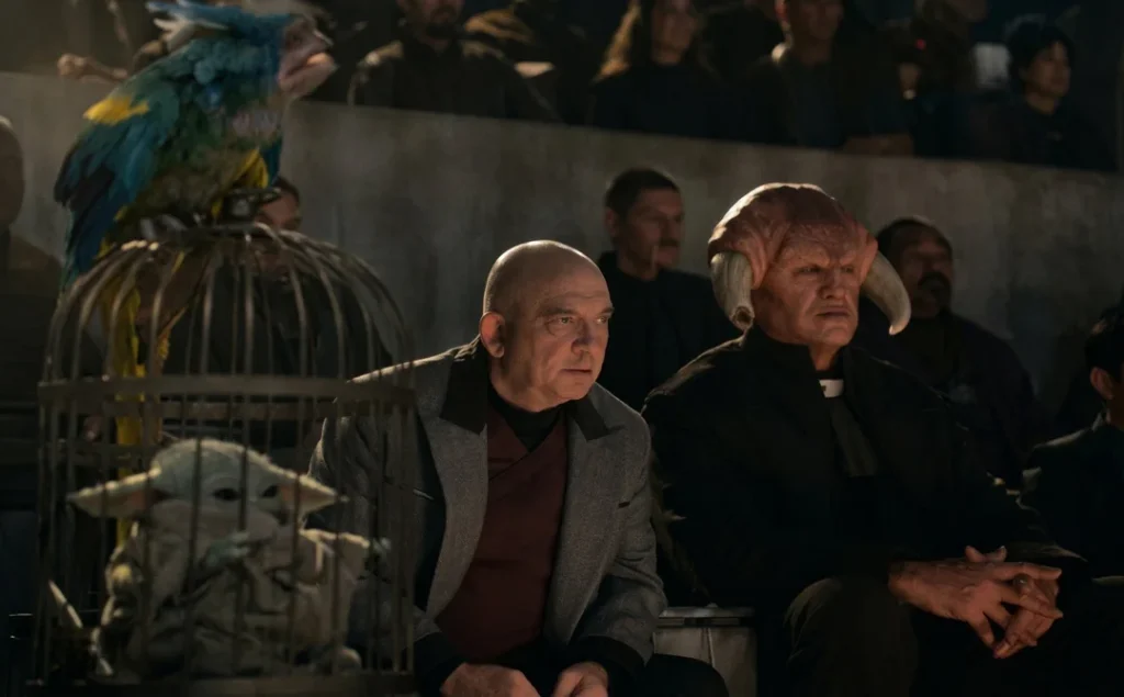 Grogu imprisoned in a birdcage next to Imperial warlord watching some entertainment with an Iktotchi