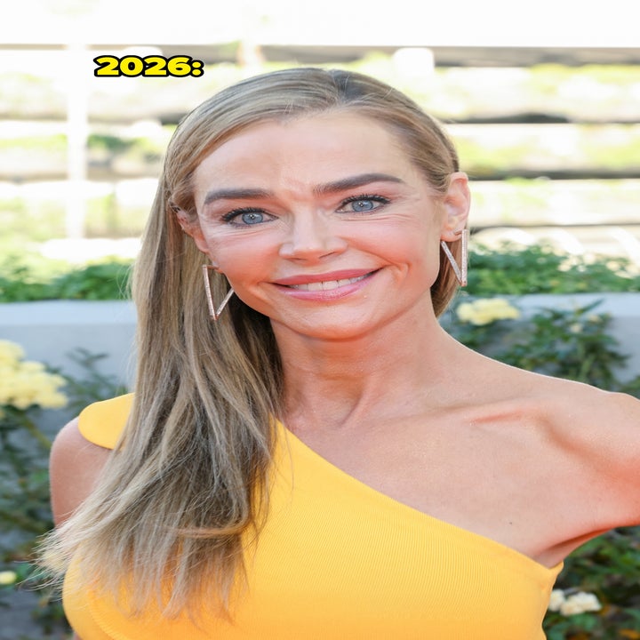 Denise Richards on the red carpet wearing an elegant one-shoulder dress, smiling at the camera with wavy long hair and geometric earrings