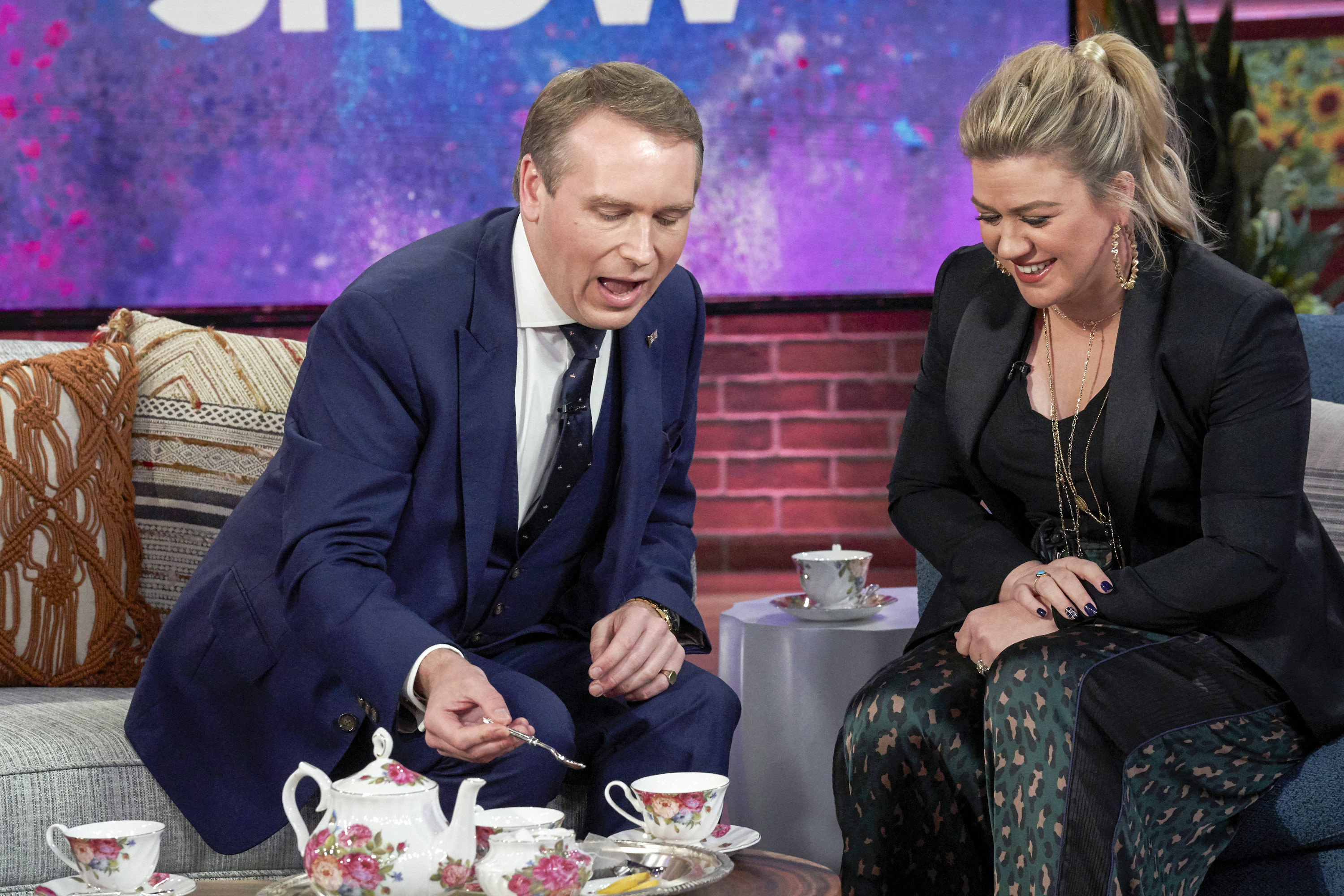 Grant Harrold on a couch stirring tea next to Kelly Clarkson