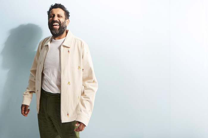 RT_Adeel Akhtar_13_09_2426284 copy Adeel Akhtar laughing as he stands in front of a light blue background.
