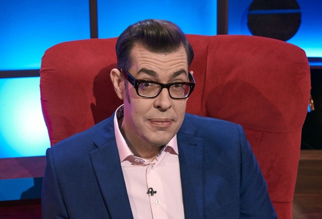 Richard Osman sits in a red chair in a blue jacket.