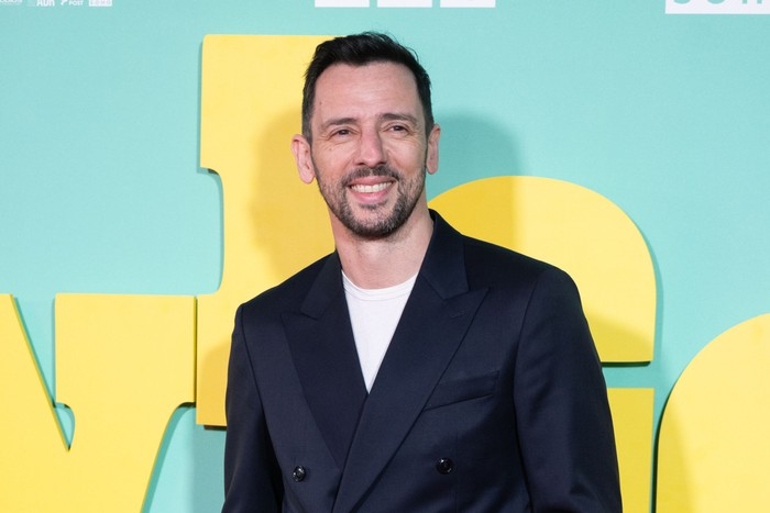 Ralf Little stands smiling ahead wearing a double breasted black suit with a white t-shirt underneath.