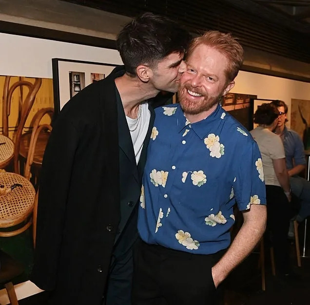 Two people share a moment, one kisses the other's cheek. The person being kissed wears a floral shirt, jeans, and has a joyful expression
