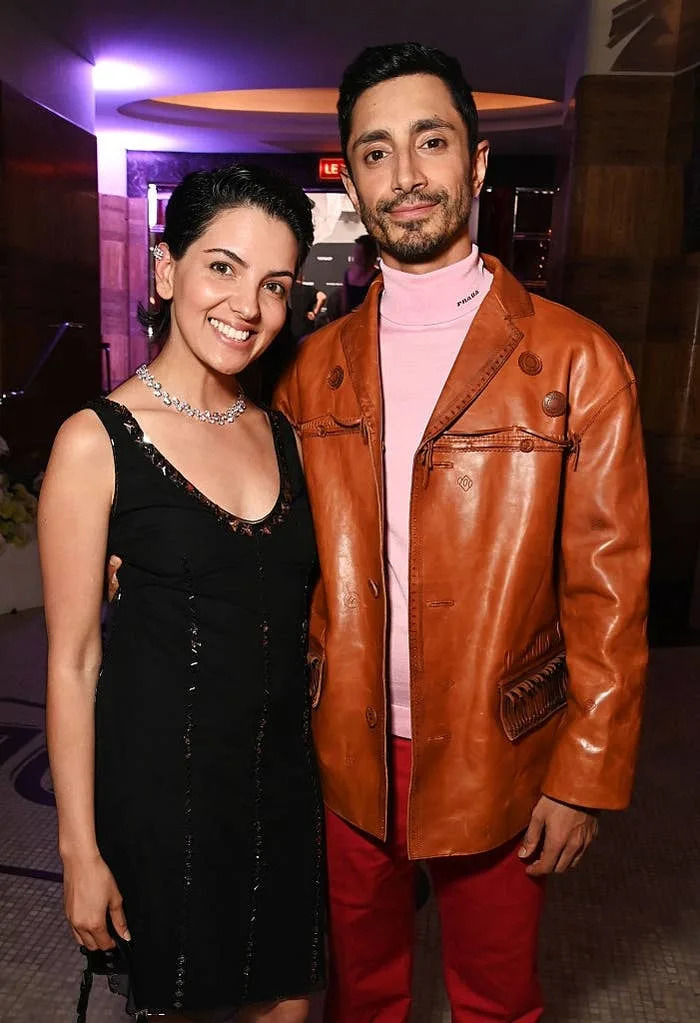Two people smiling at a formal event; one in a black dress, the other in a brown leather jacket and pink turtleneck