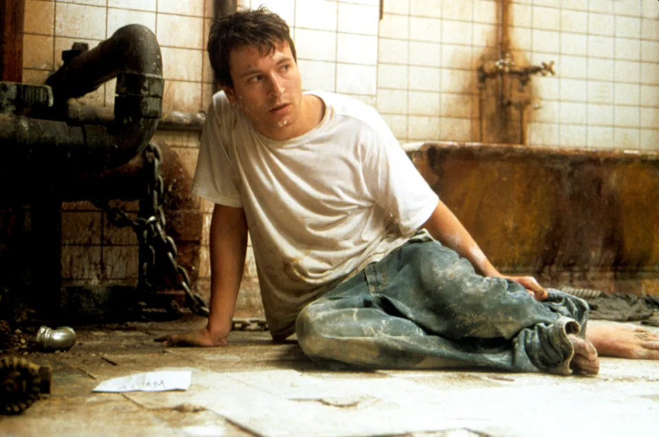 A scene from a movie showing a distressed man in a dirty room, sitting on the floor, wearing a worn-out shirt and jeans, looking at something off-camera