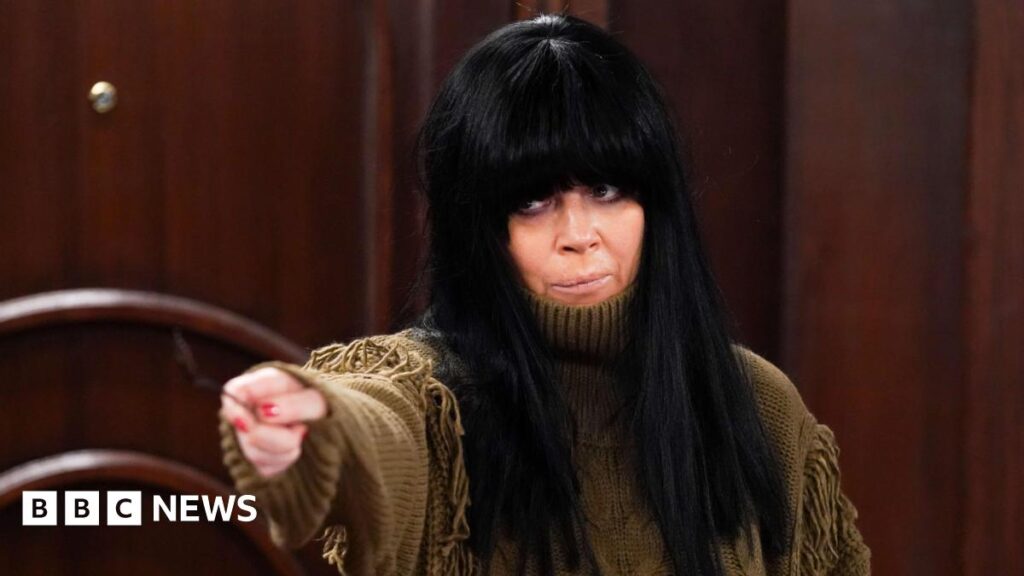 Claudia Winkleman played by Anna Friel, who is wearing a brown fringed jumper and a black wig with a blunt fringe.